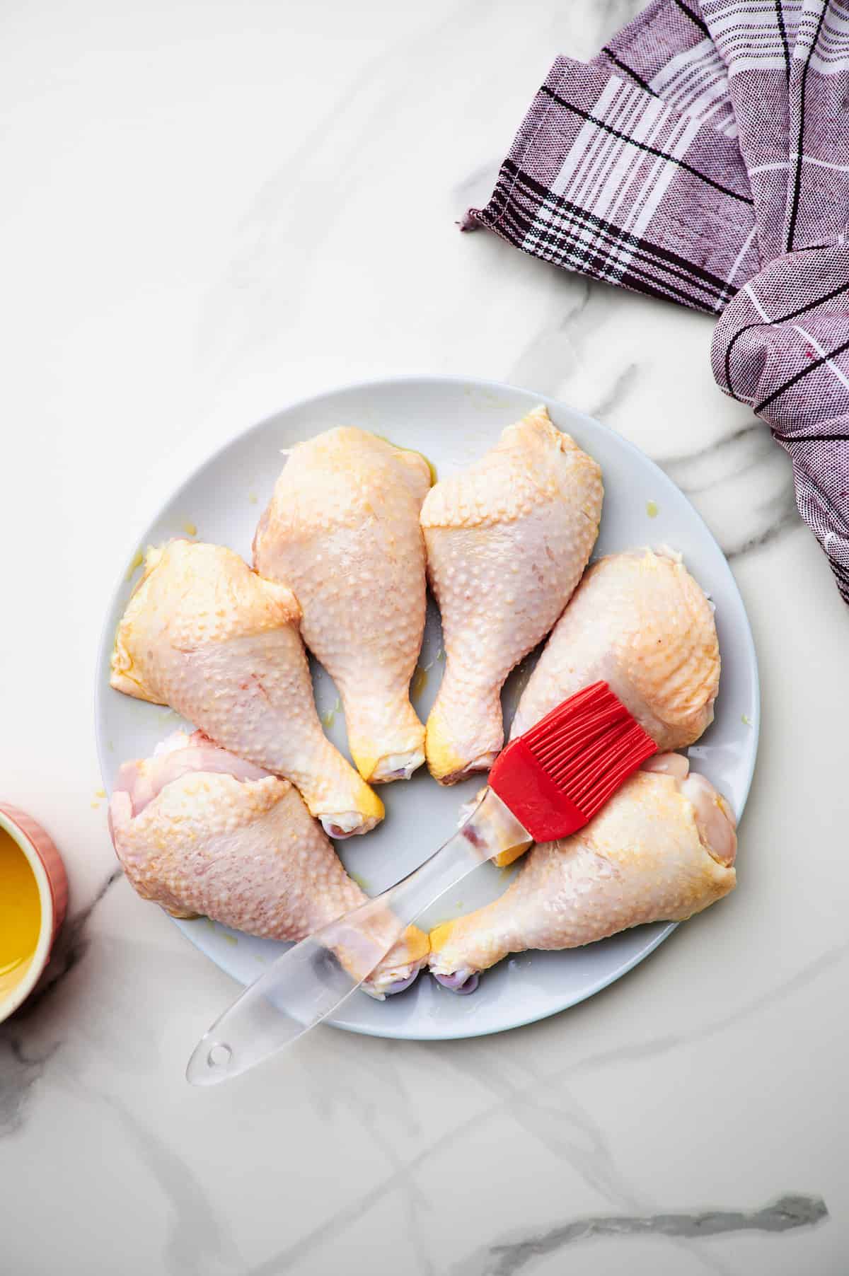 Brushing  uncooked drumsticks with olive oil on a white plate.