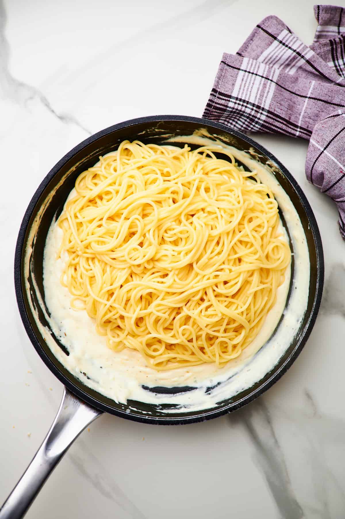 adding parmesan to a skillet and tossing pasta with creamy garlic sauce