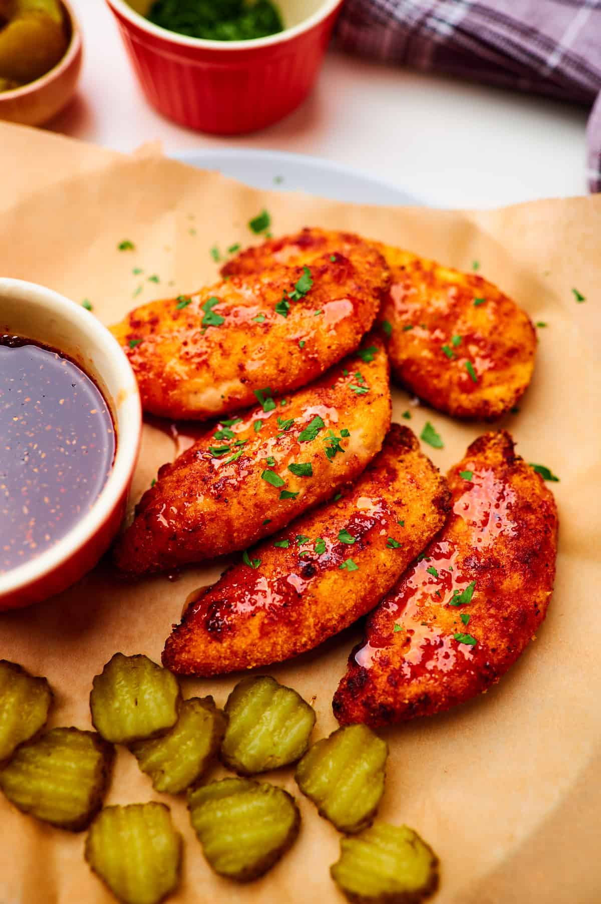 pile of air fryer chicken tenders drizzled in homemade hot honey sauce and sprinkled with parsley, served with a side of sliced pickles in a small ramekin.