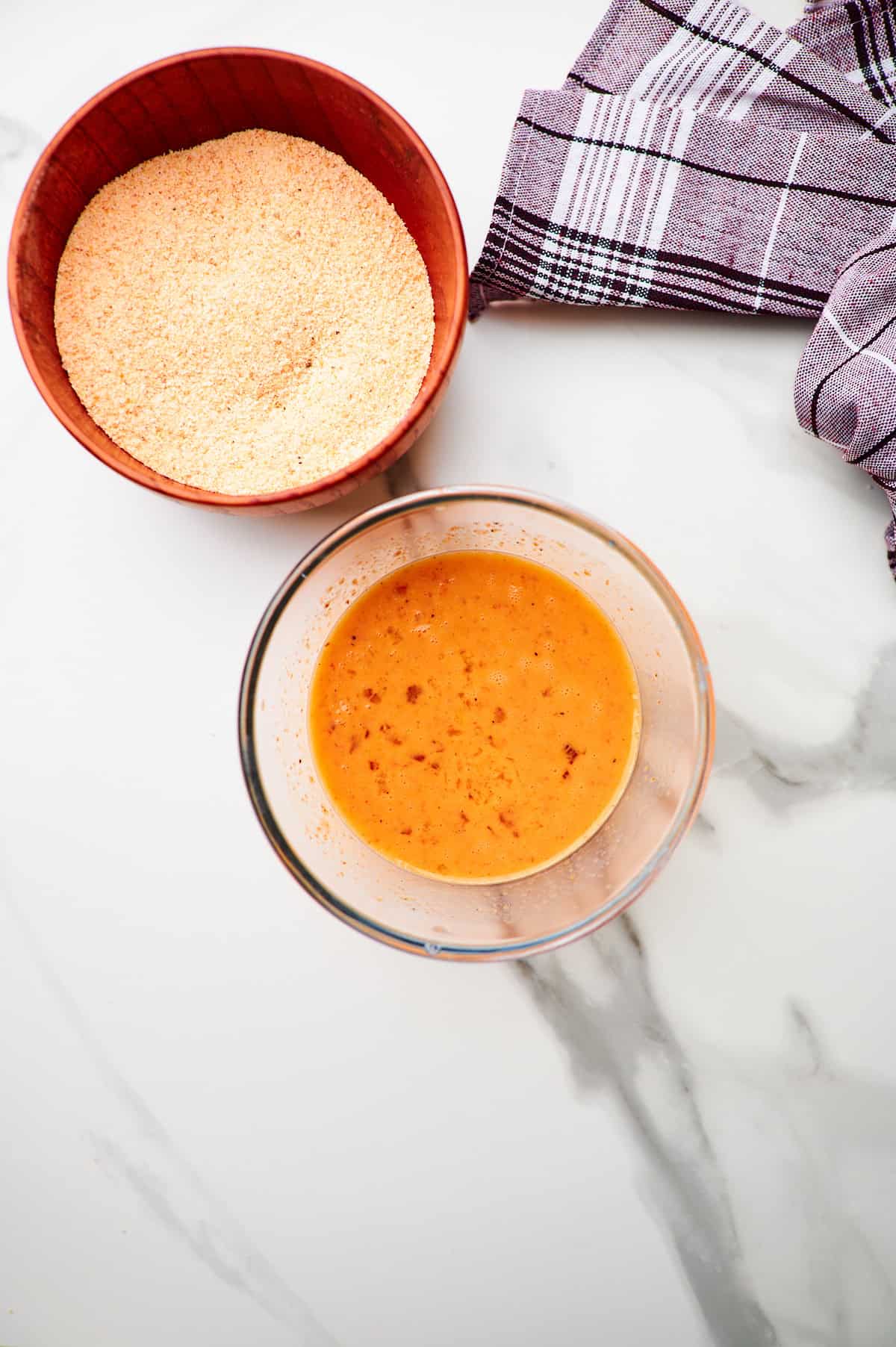 whipped eggs in a bowl with milk, hot sauce, and seasoning added in the center of the frame and the breadcrumb mixture in a separate bowl in the top of the frame.