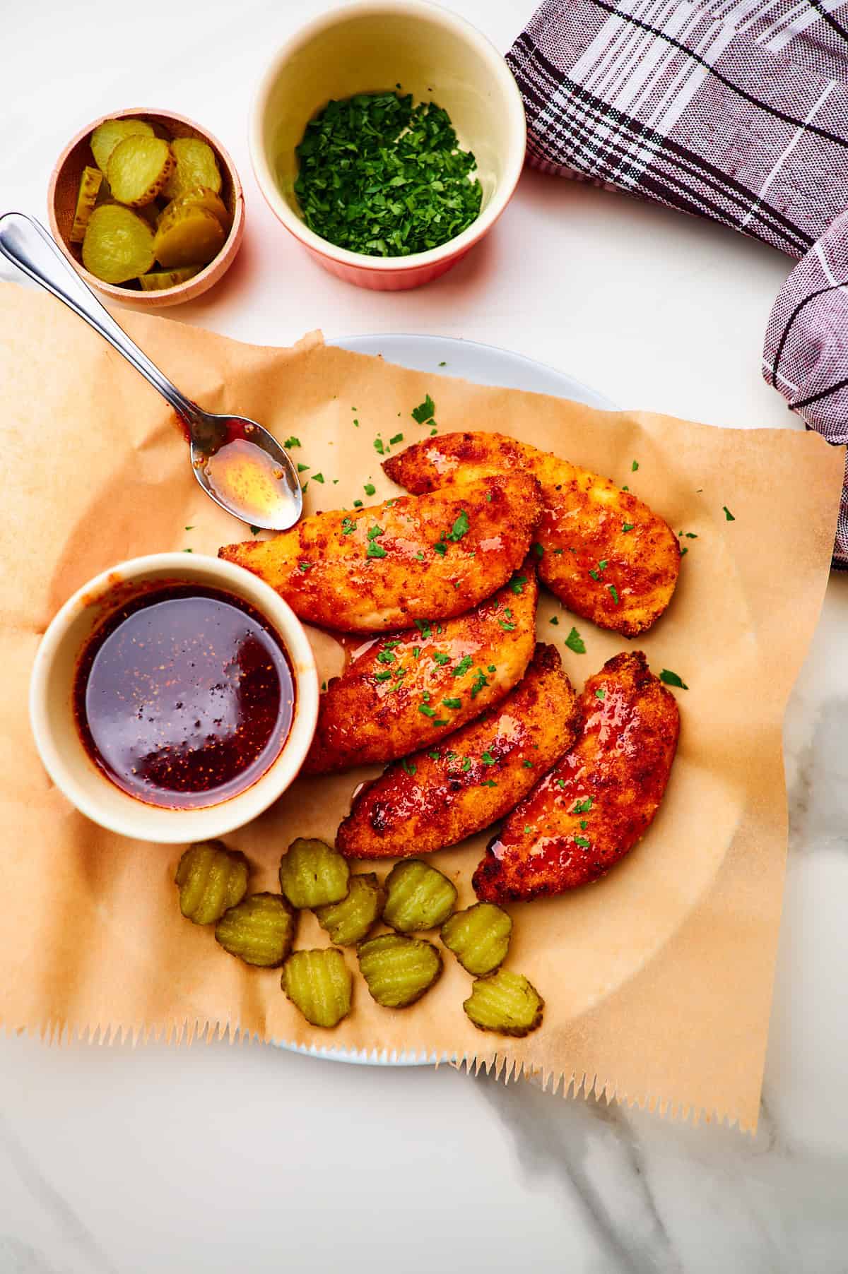 round white dinner plate topped with a crumpled piece of white parchment paper with honey hot chicken tenders drizzled with hot honey sauce, sprinkled with parsley, and served with sliced dill pickles with extra sauce and extra pickle slices in small bowls in the top and bottom of the frame.