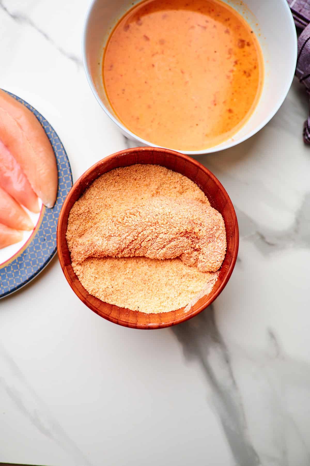 dredging station set up with a single chicken tender that had already been dipped into the egg wash bowl at the top of the frame covered in the breadcrumb mixture in the middle of the frame with a plate of raw, naked tenders in the very bottom corner of the frame.