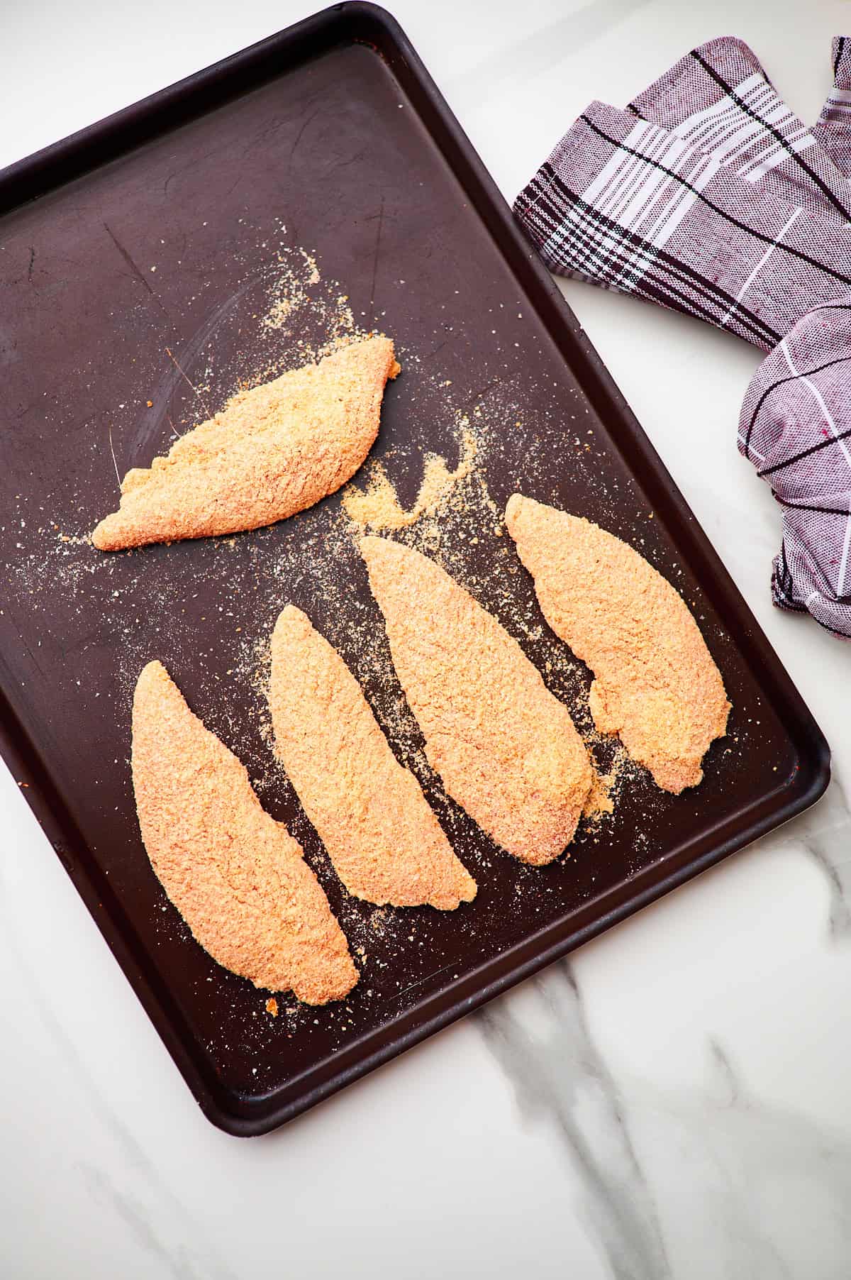 silver baking tray with 12 homemade breaded chicken tenders that have yet to be cooked.