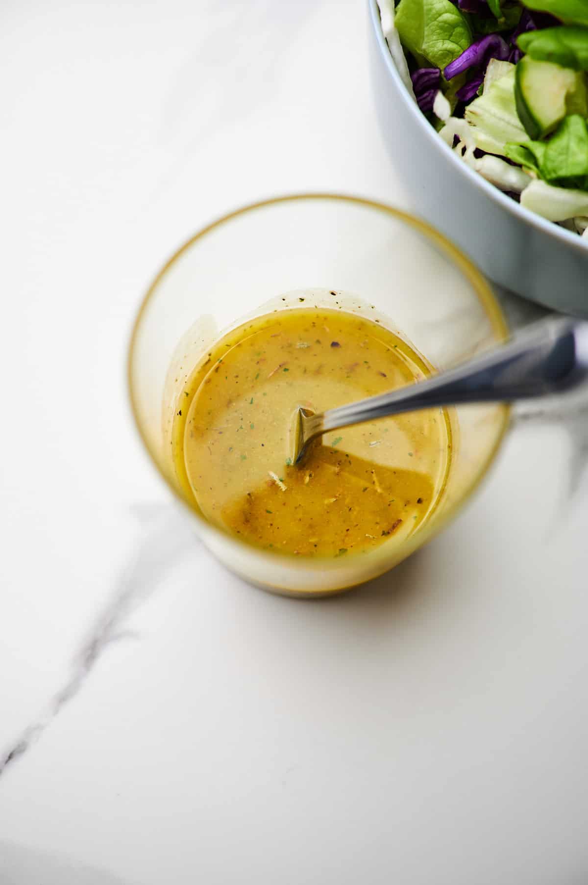 A small glass jar of italian submarine dressing with a spoon and a salad.