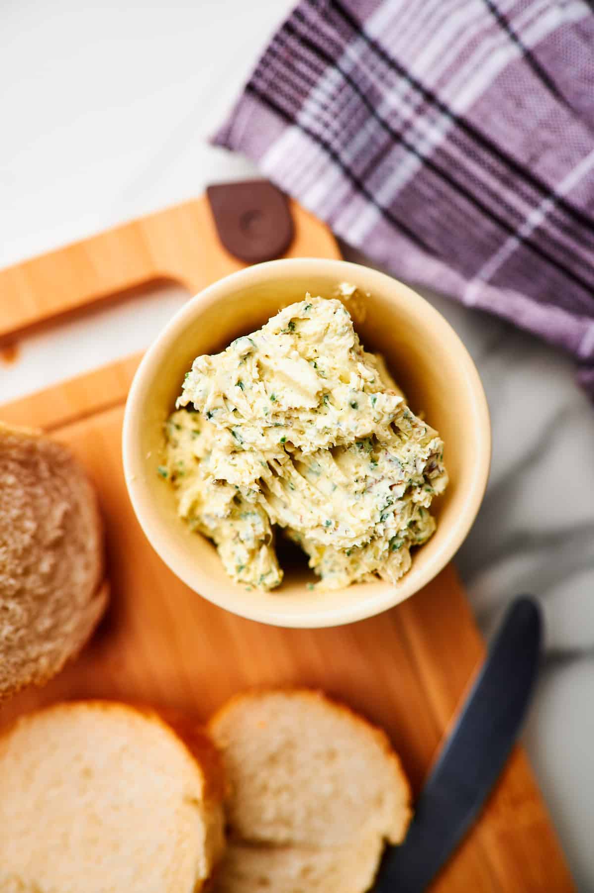 bowl of roasted garlic butter with seasoning and herbs on cutting board with bread and rosemary.